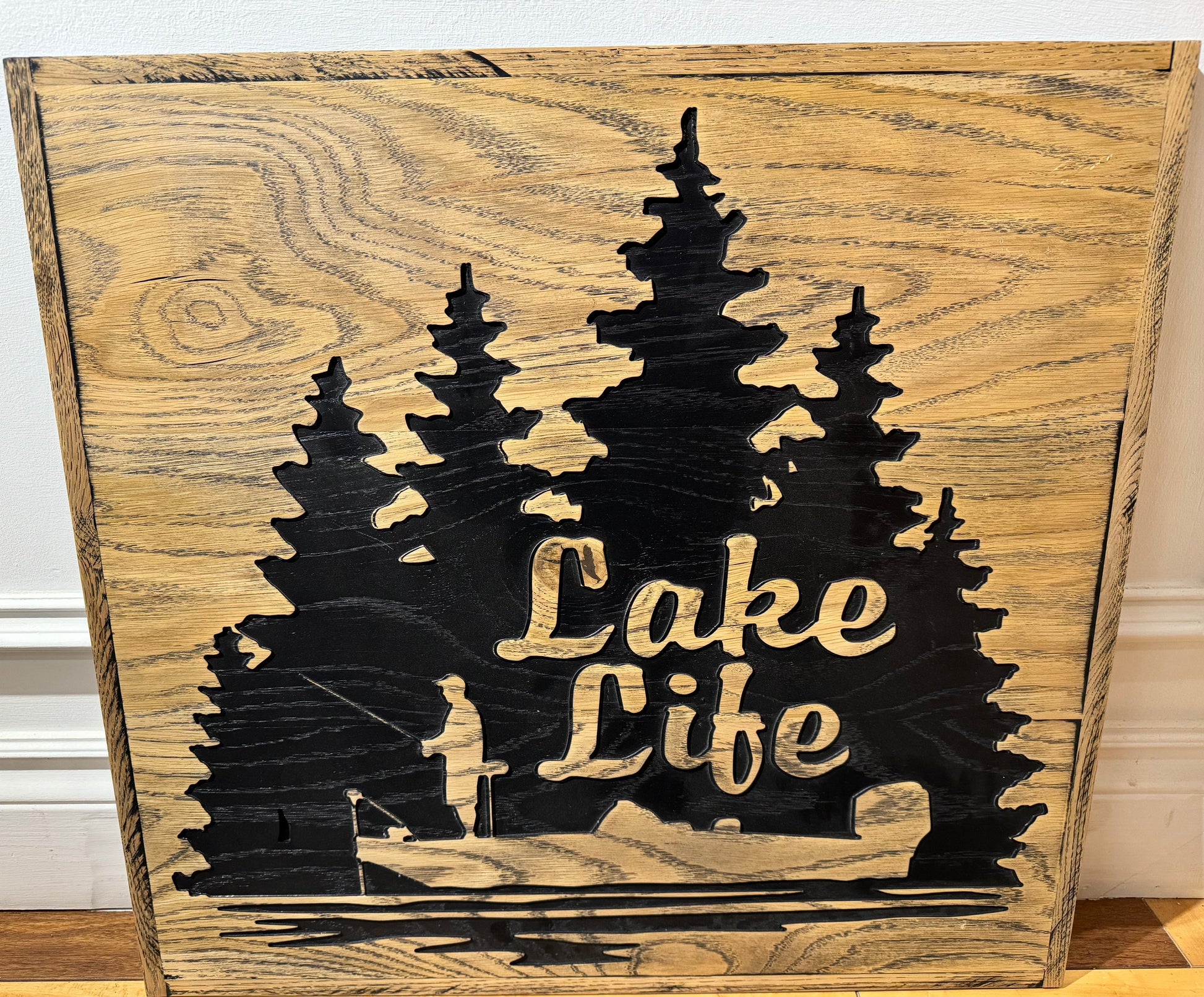 Wooden sign of man standing in a small boat fishing. Lake life is spelt in the shadows of the trees behind the fisherman.